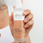 Hand holding a bottle of Glow Skin Beauty Barrier Body Serum against a neutral background