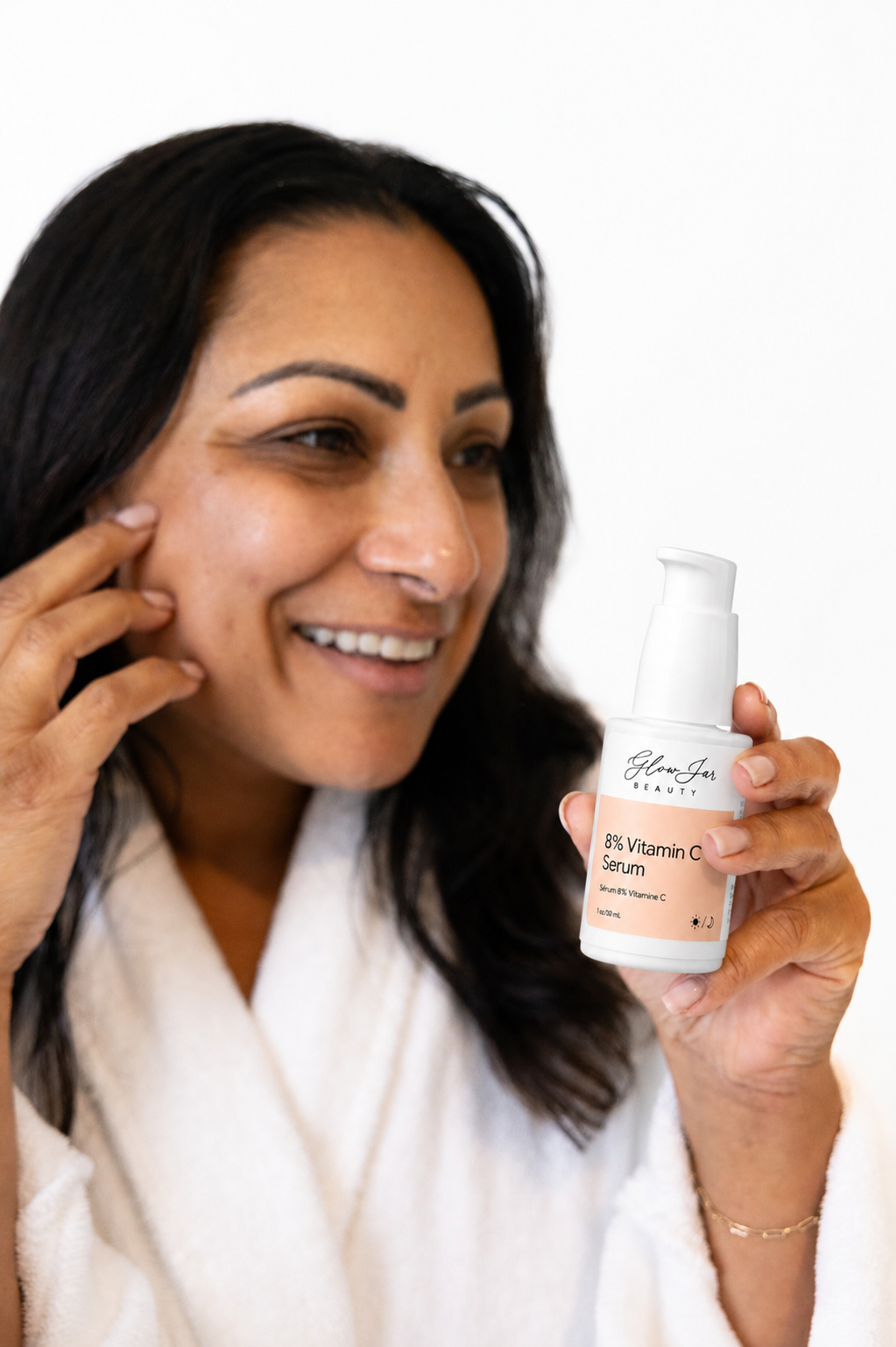 A woman holds up a bottle of Glow jar Beauty 8% Vitamin C Serum, smiling and touching her cheek against a clean white background.