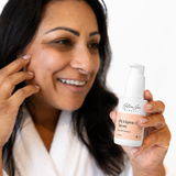 A woman holds up a bottle of Glow jar Beauty 8% Vitamin C Serum, smiling and touching her cheek against a clean white background.