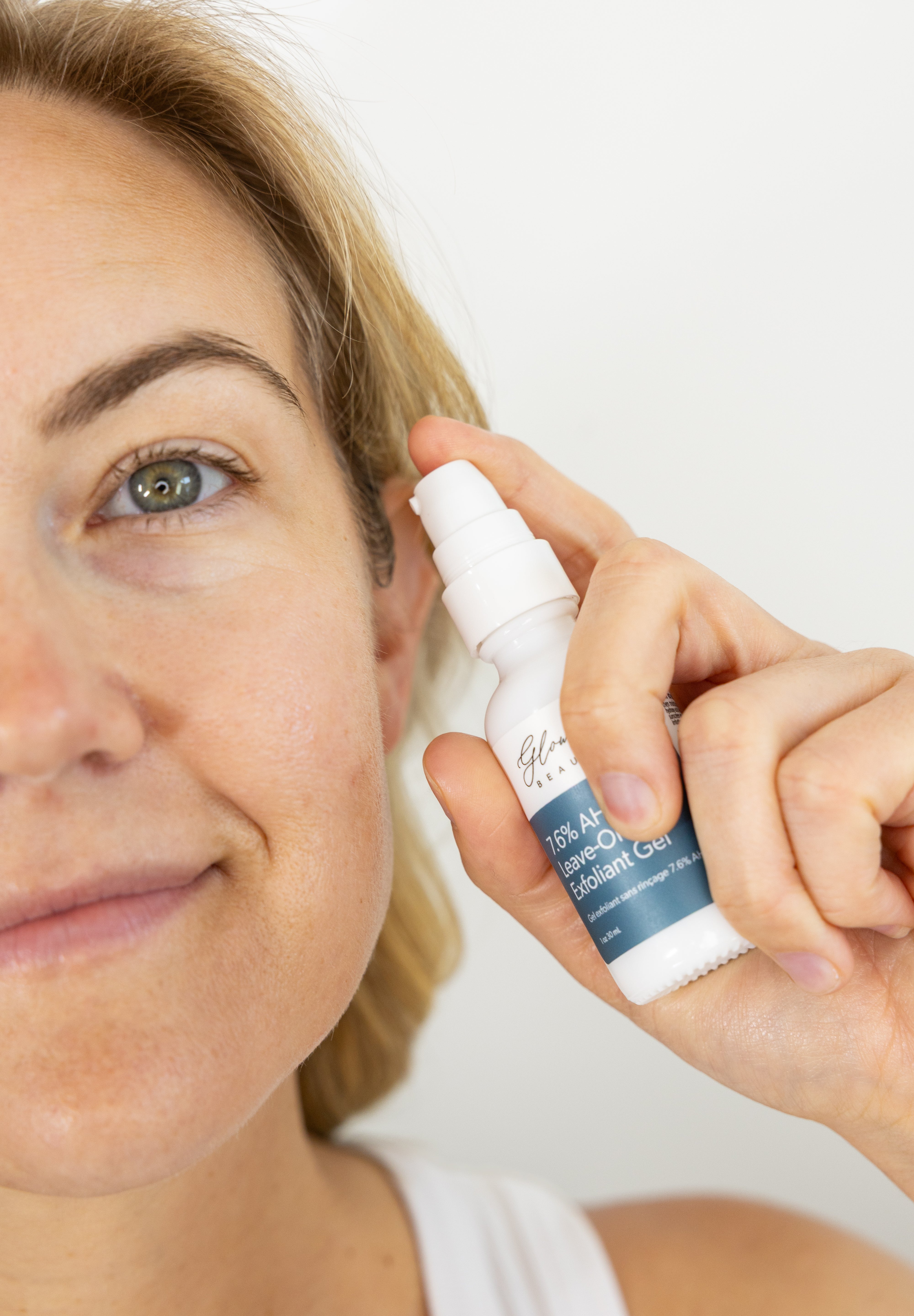 Woman applying 7.6% AHA Leave On Exfoliant skincare product to her face with a white background