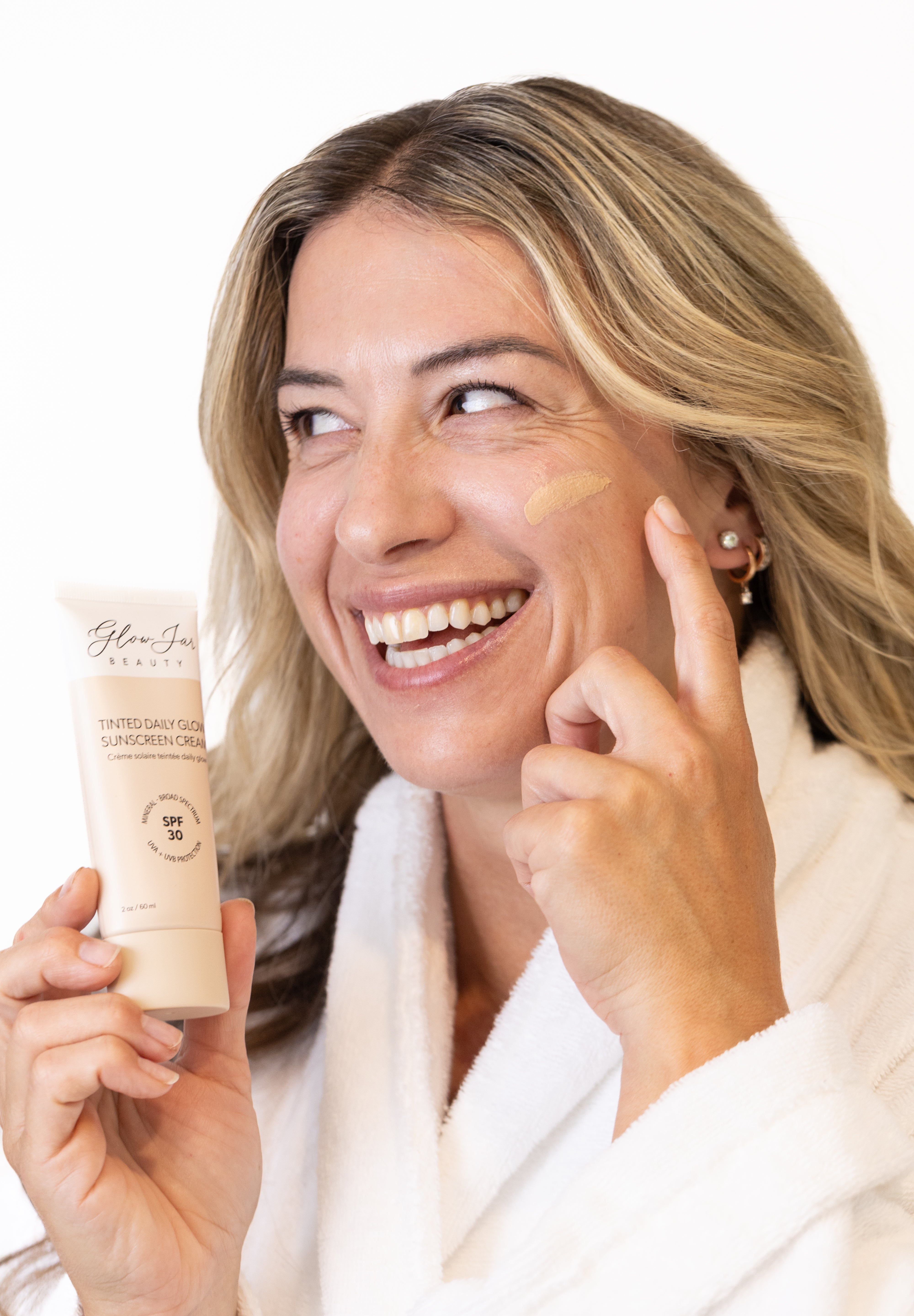 Woman applying Glow jar Beauty tinted SPF sunscreen cream to her face with a tube of sunscreen in hand.