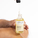 Hand holding a bottle of pineapple body oil with a dropper, on a white background.