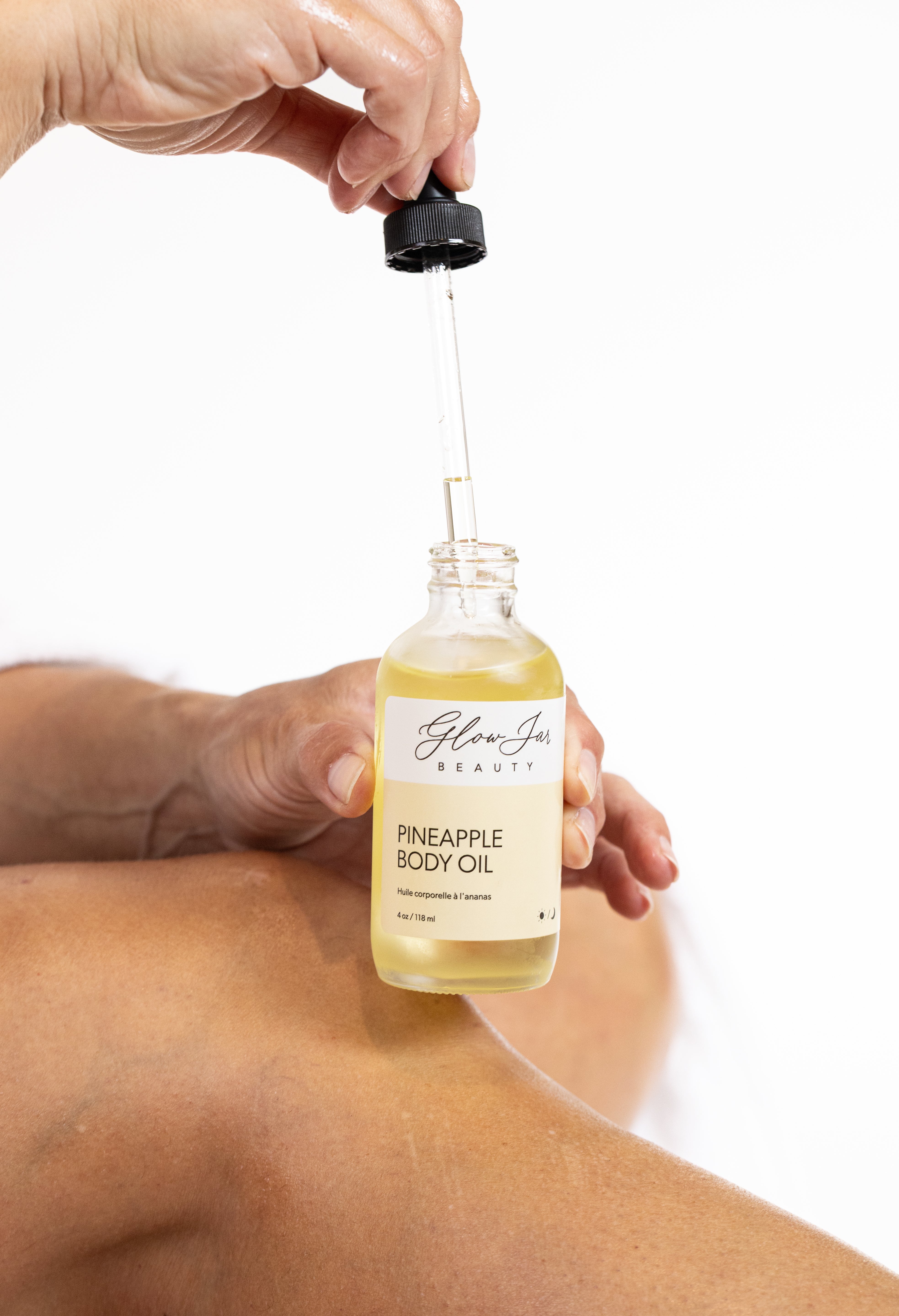 Hand holding a bottle of pineapple body oil with a dropper, on a white background.